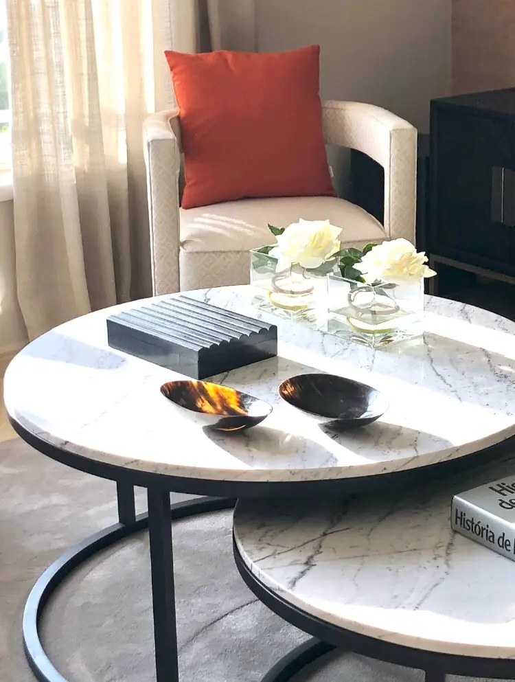 A large armchair upholstered in beige fabric, stone coffee tables decorated with flowers, and bone boxes and bowls