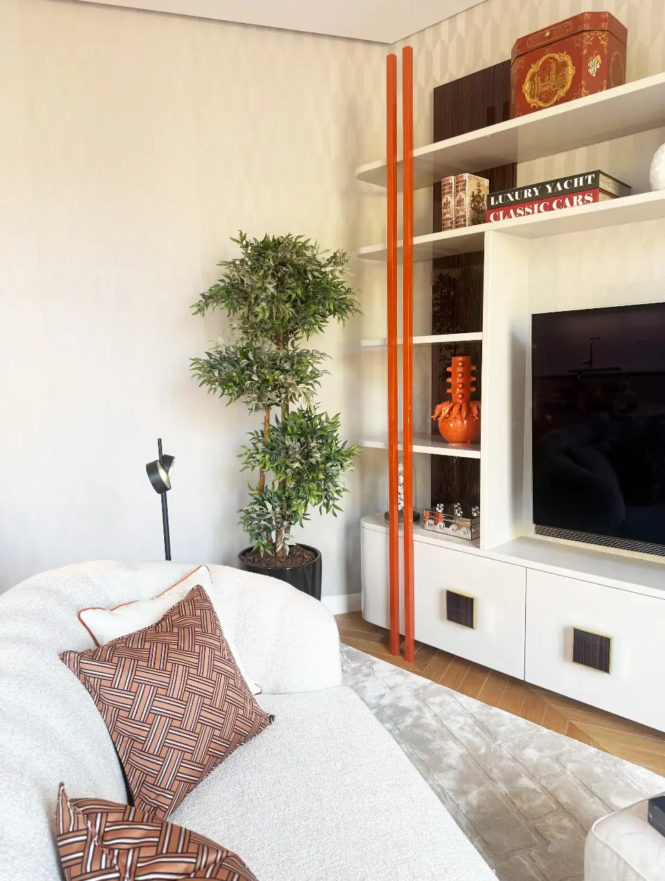Sala revestida a papel de parede geométrico com estante TV lacada beige, sofá branco com almofadas decorativas laranja