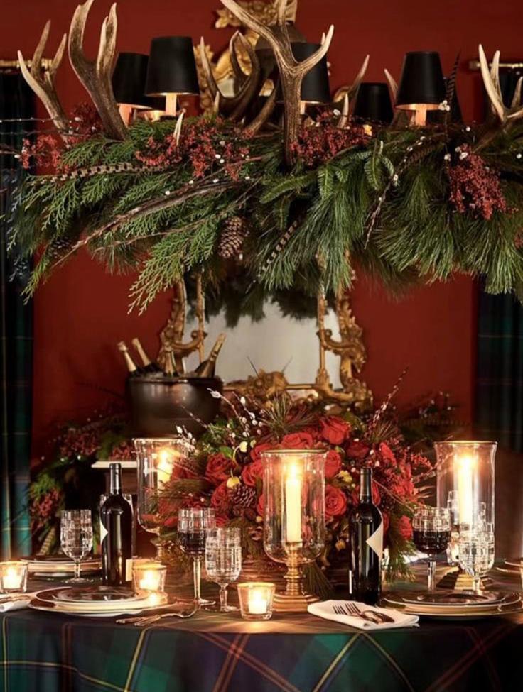 Decoração de mesa de natal com candeeiro pendente coberto de pinheiros e azevinhos. No centro da mesa está um arranjo floral em tons de encarnado