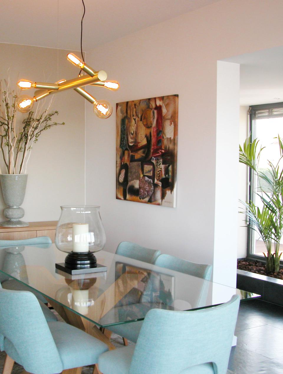 Decoração de sala de jantar com cadeiras estofadas em tecido verde água e mesa de jantar em vidro. Ao fundo arranjo de ramagens e palmeira natural