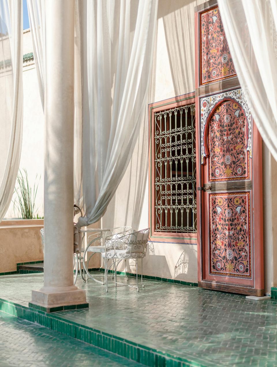 Pátio com pavimento em mosaico zellige verde, porta encarnada decorada e cortinas brancas