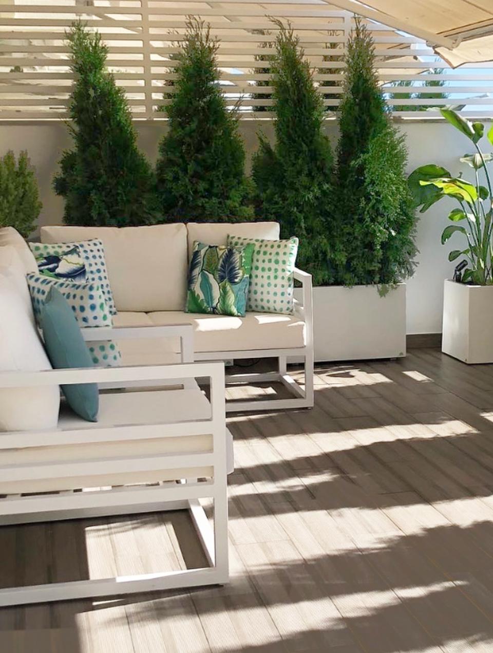 Terrace with brown ceramic tiles on the floor, white sofas with decorative cushions in shades of blue and green, and pots with cedar plants