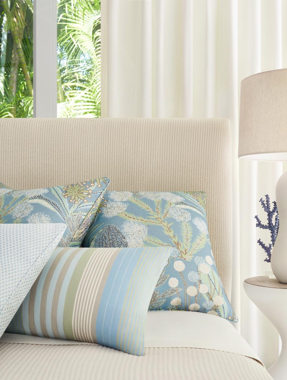 Cama com cabaceira beige e almofadas decorativas azuis com padrão floral, almofadas de riscas em tons de verde e azul e leve cortina branca ao fundo