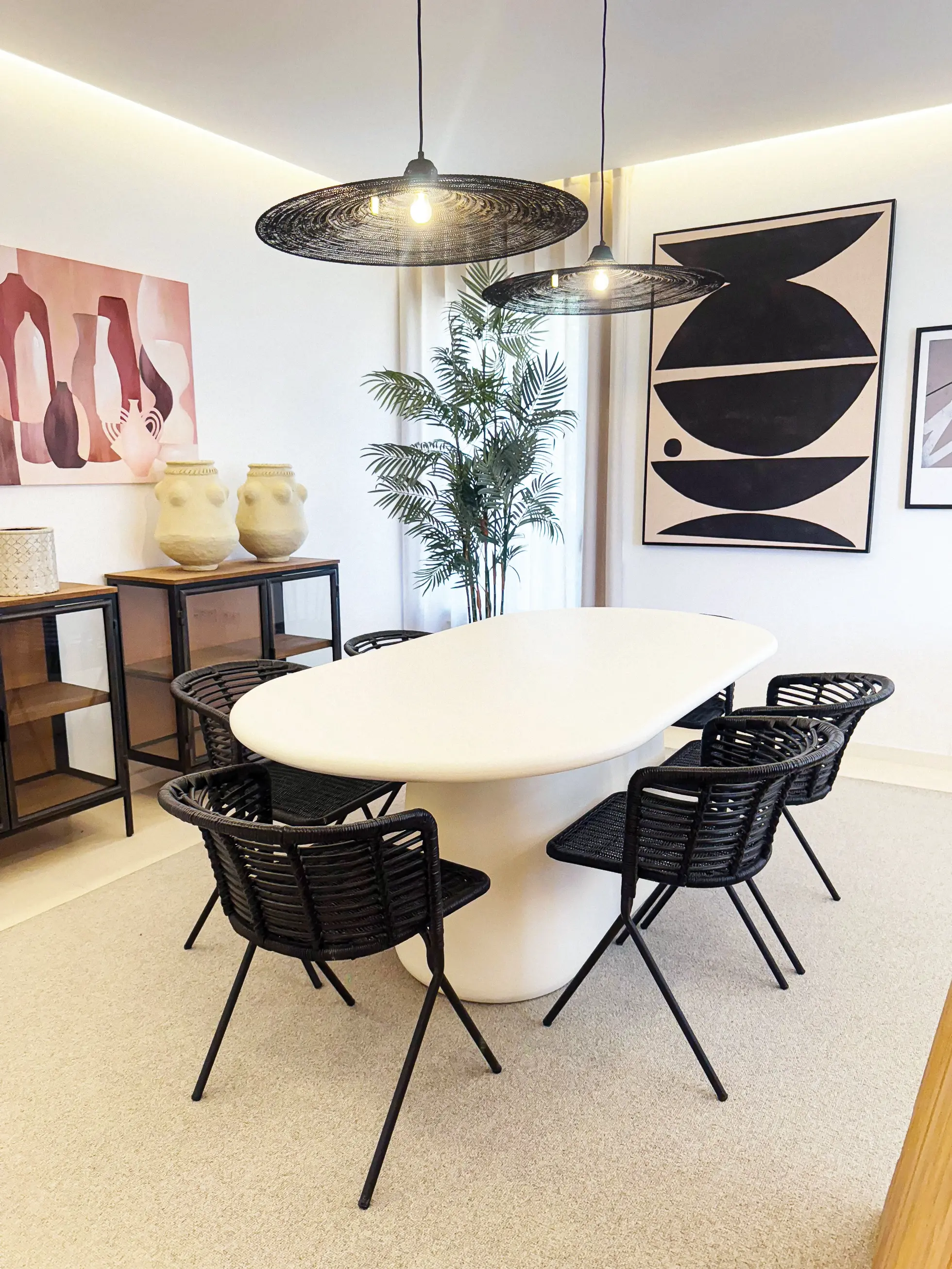 Sala de jantar com mesa branca, cadeiras de ratan preto, aparadores em madeira com portas de vidro e metal e quadro beige e preto ao fundo