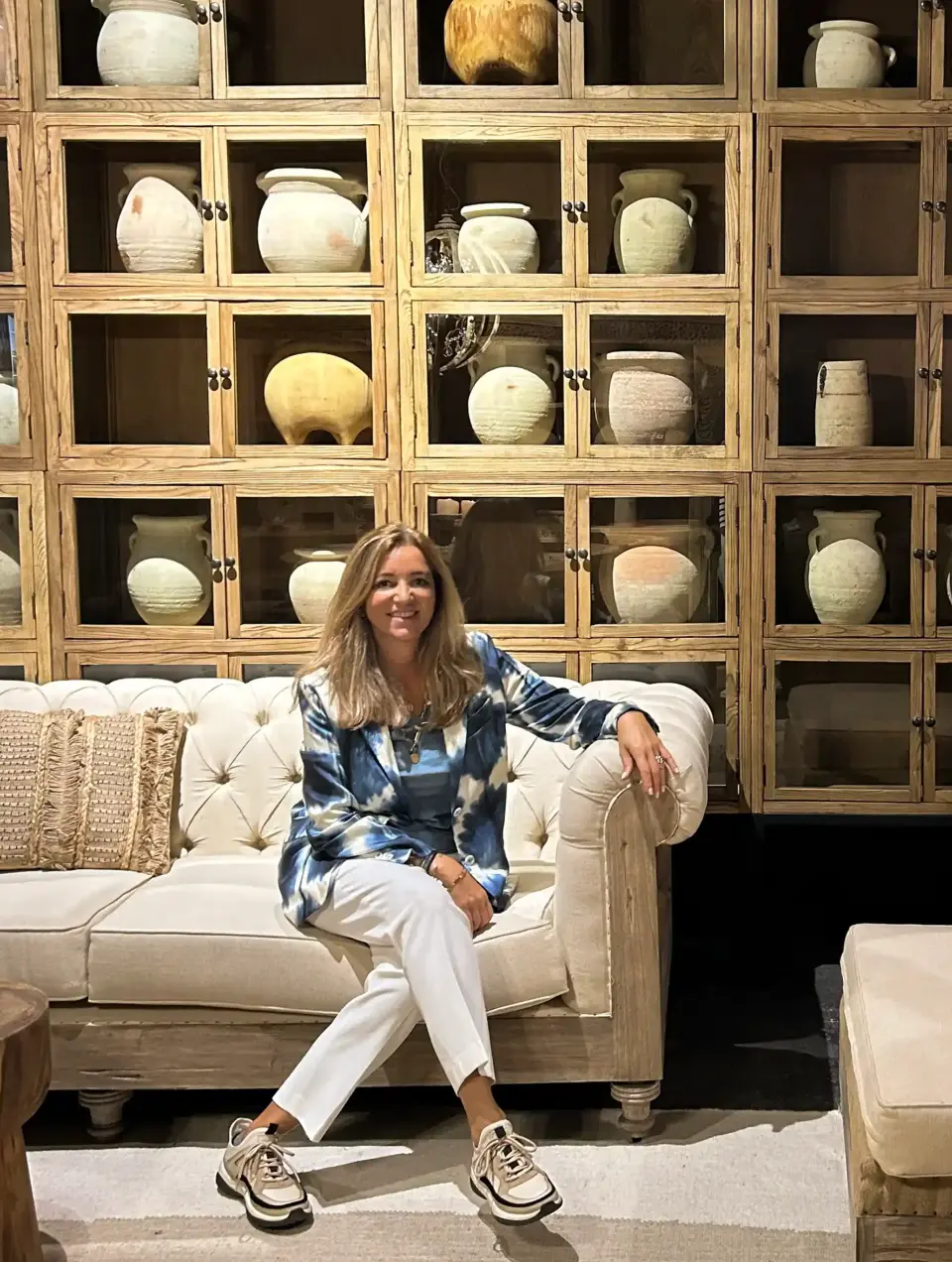 Arquiteta de Interiores Margarida Bugarim sentada num sofá branco, ao fundo uma estante de madeira cheia de vasos e potes antigos