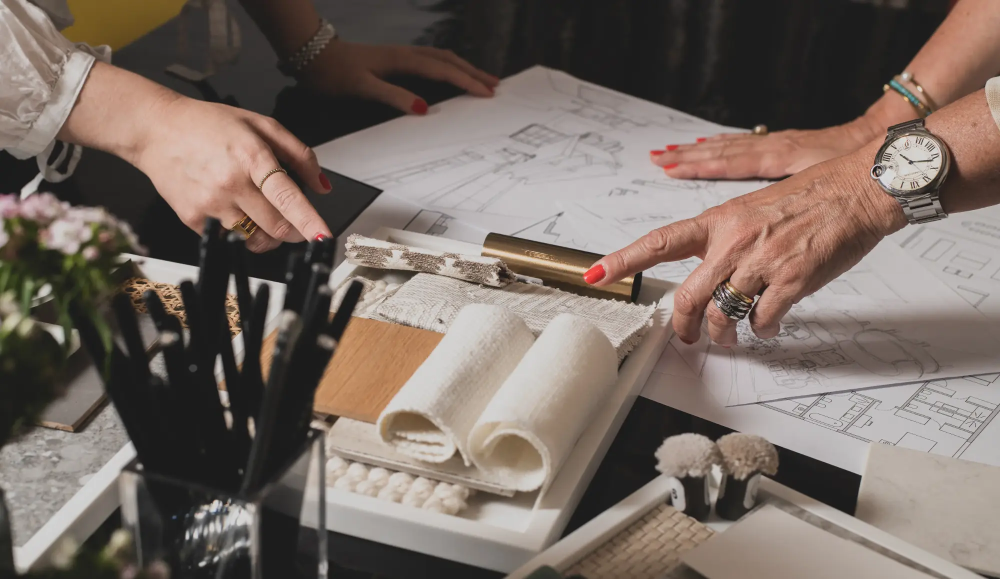 Mãos de designers de interiores a analisar amostras de tecidos, materiais para projeto em desenvolvimento com plantas e alçados sobre a mesa.
