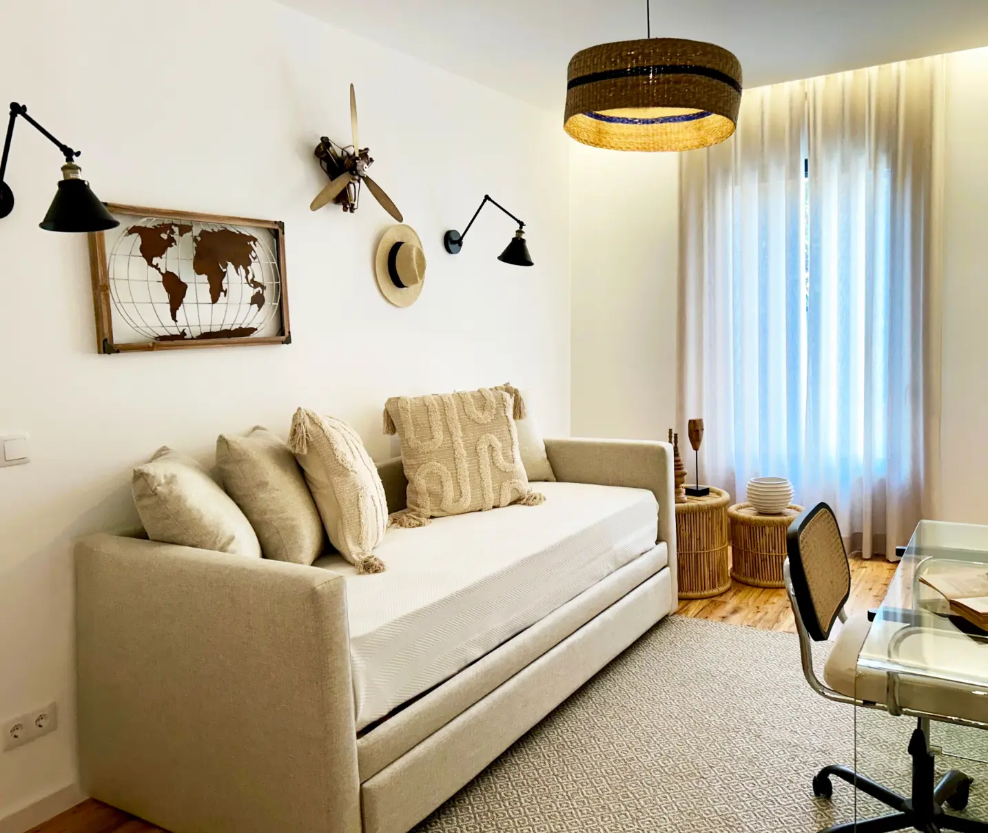 Decoração de escritório com Cama-nido efeito sofá em tons neutos, secretária de vidro e cadeira em palhinha, decoração de parede alusiva a viagens