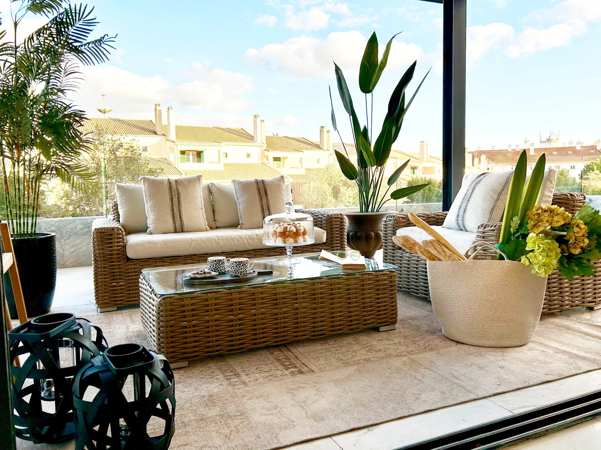 Decoração de terraço com sofás e mesa em ratan sintético castanho, almofadas beiges, plantas tropicais e lanternas pretas