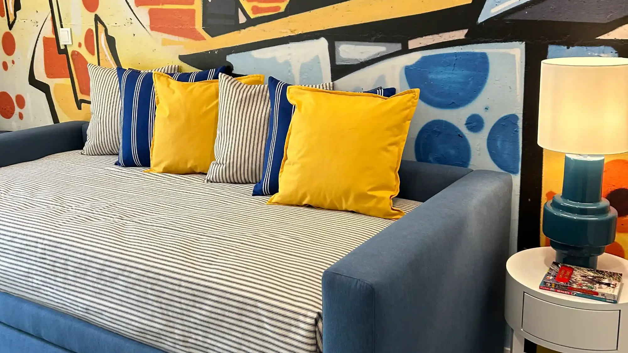 Quarto jovem com mural de grafitty na cabeceira da cama, uma cama dupla tipo sofá e decoração em tons de azul e amarelo.