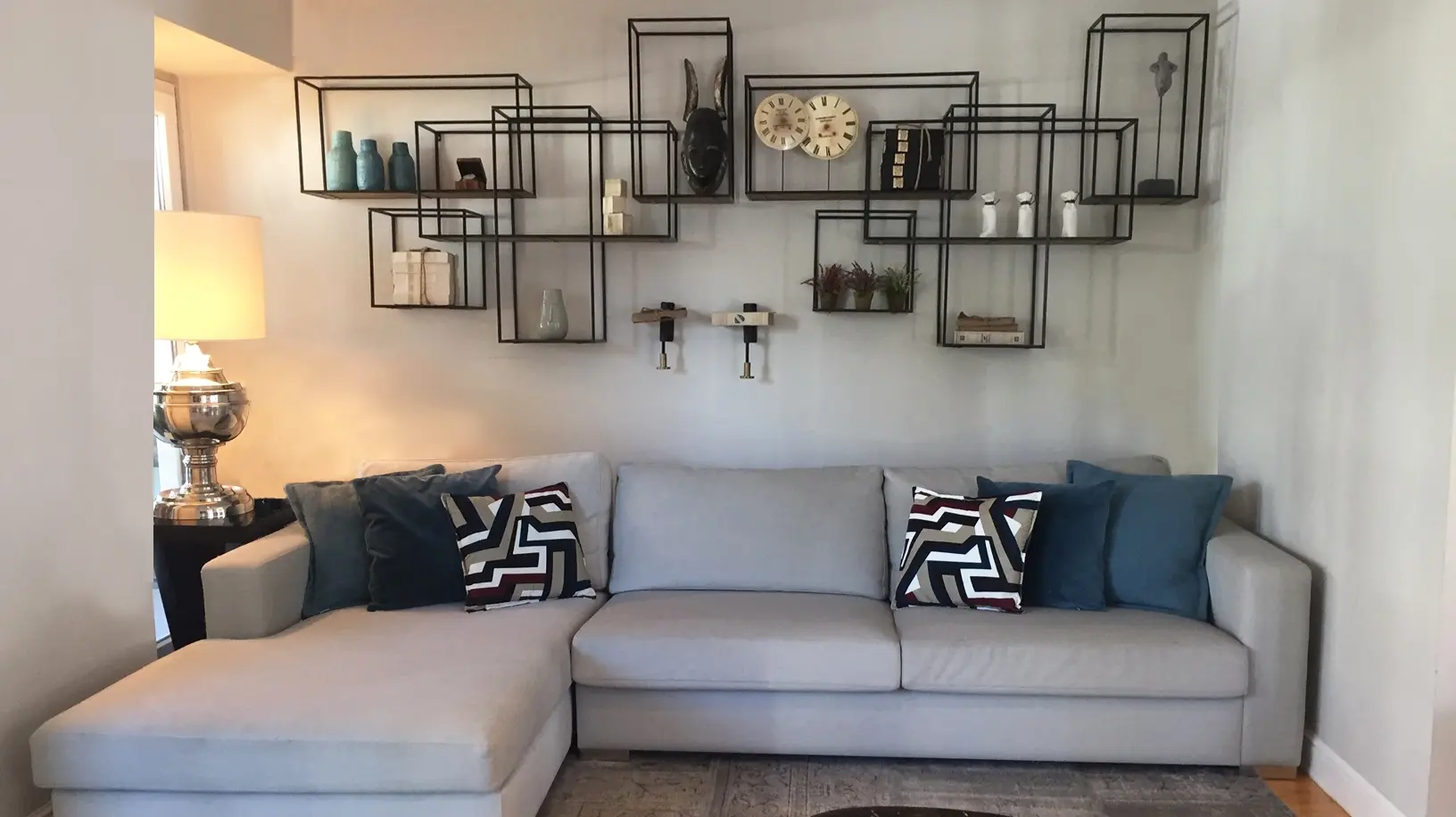 Sofá cinzento com chaise-longue e almofadas decorativas em tons de azul. Sobre o sofá estantes de parede industriais com peças decorativas variadas
