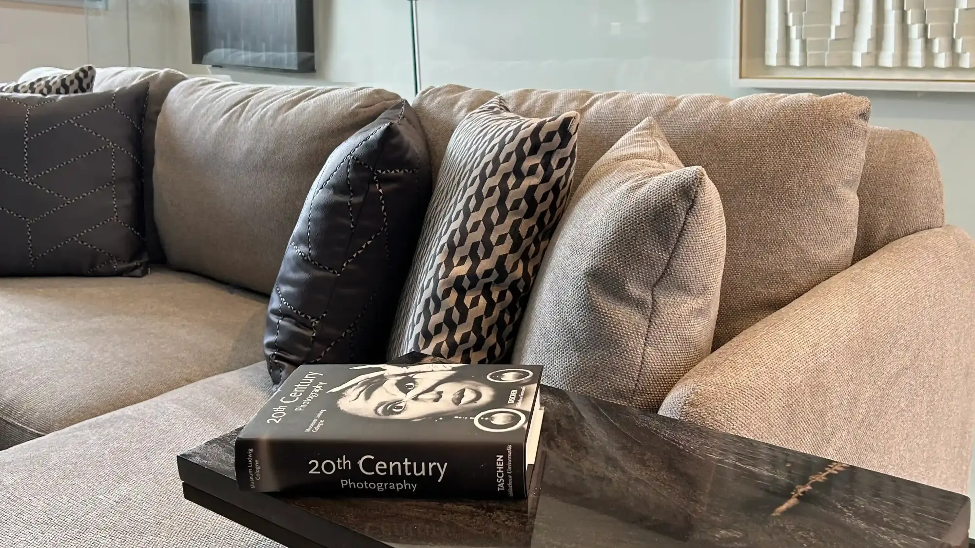 Sala de estar com sofá confortável cinzento, almofadas com padrões cinza e pretos e livro sobre fotografia- decoração assinada por Margarida Bugarim